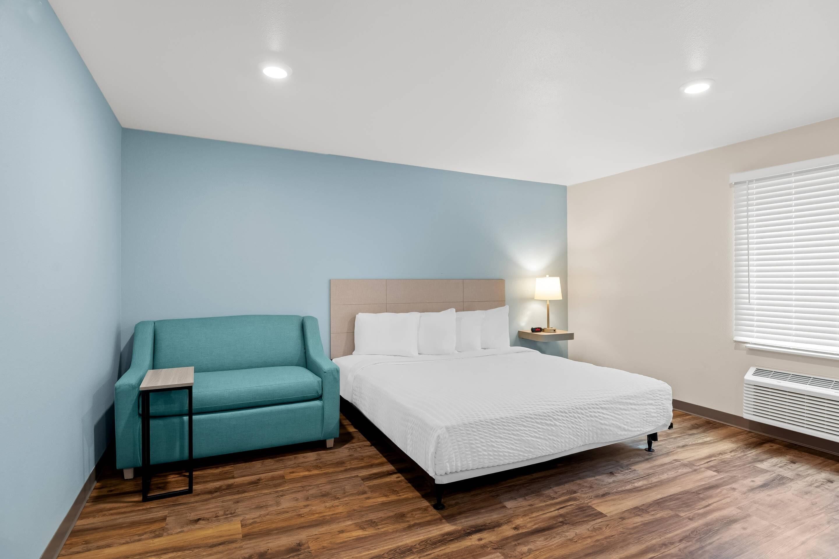 Ein Hotelzimmer mit Bett, einem blaugrünen Sofa, einem kleinen Tisch, einem Nachttisch mit Lampe und einem Fenster mit Jalousien.