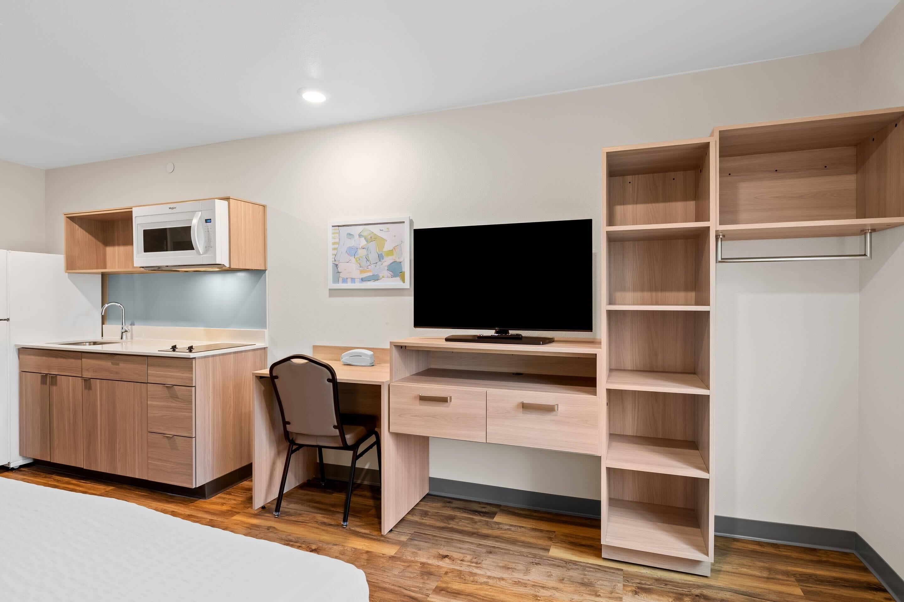A modern, compact room with a kitchenette, a desk with a computer, a television, and built-in shelving.