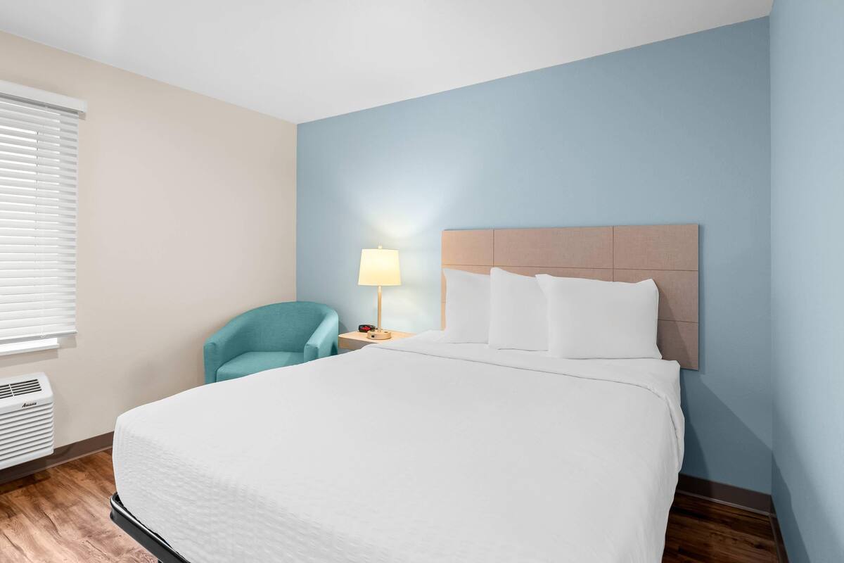 Habitación de hotel con una cama grande, una mesita de noche con lámpara, una silla y una ventana con persianas.