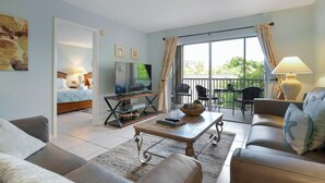 A living room with a sofa, a coffee table, and a TV. There is a balcony with chairs and a table. - Palm Bay Club Private Balcony Beach Side  (Sarasota)