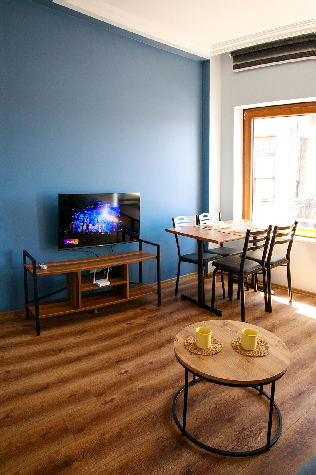 Ein Zimmer mit Fernseher auf einem Holzbalken, einem Esstisch mit Stühlen und einem kleinen runden Tisch mit zwei gelben Tassen.