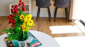 A modern room with a white round table, two dark chairs, a TV mounted on the wall, a window with sheer curtains, and a vase with flowers on the table.