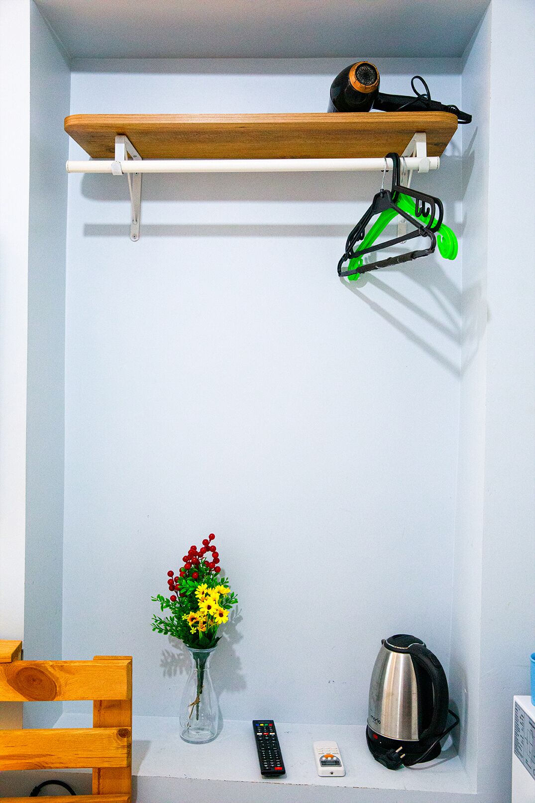 Een kleine kamer met een houten plank, een vaas met bloemen, een waterkoker en twee afstandsbedieningen.
