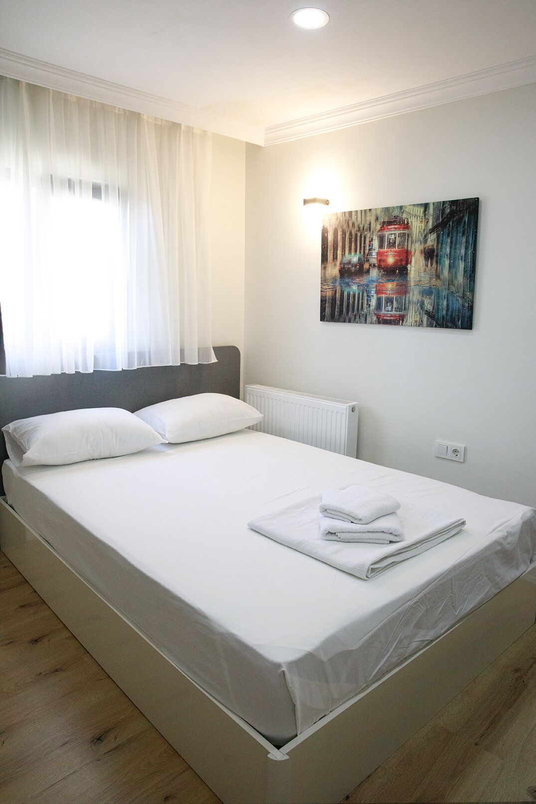 A neatly made bed with white linens and a folded towel, a radiator, and a painting on the wall.