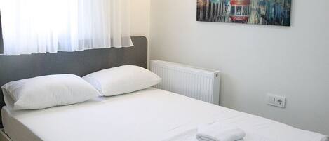 A neatly made bed with white linens and a folded towel, a radiator, and a painting on the wall.