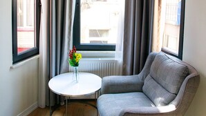 A corner room with a grey armchair, a small round table with a vase of flowers, and a window with curtains.