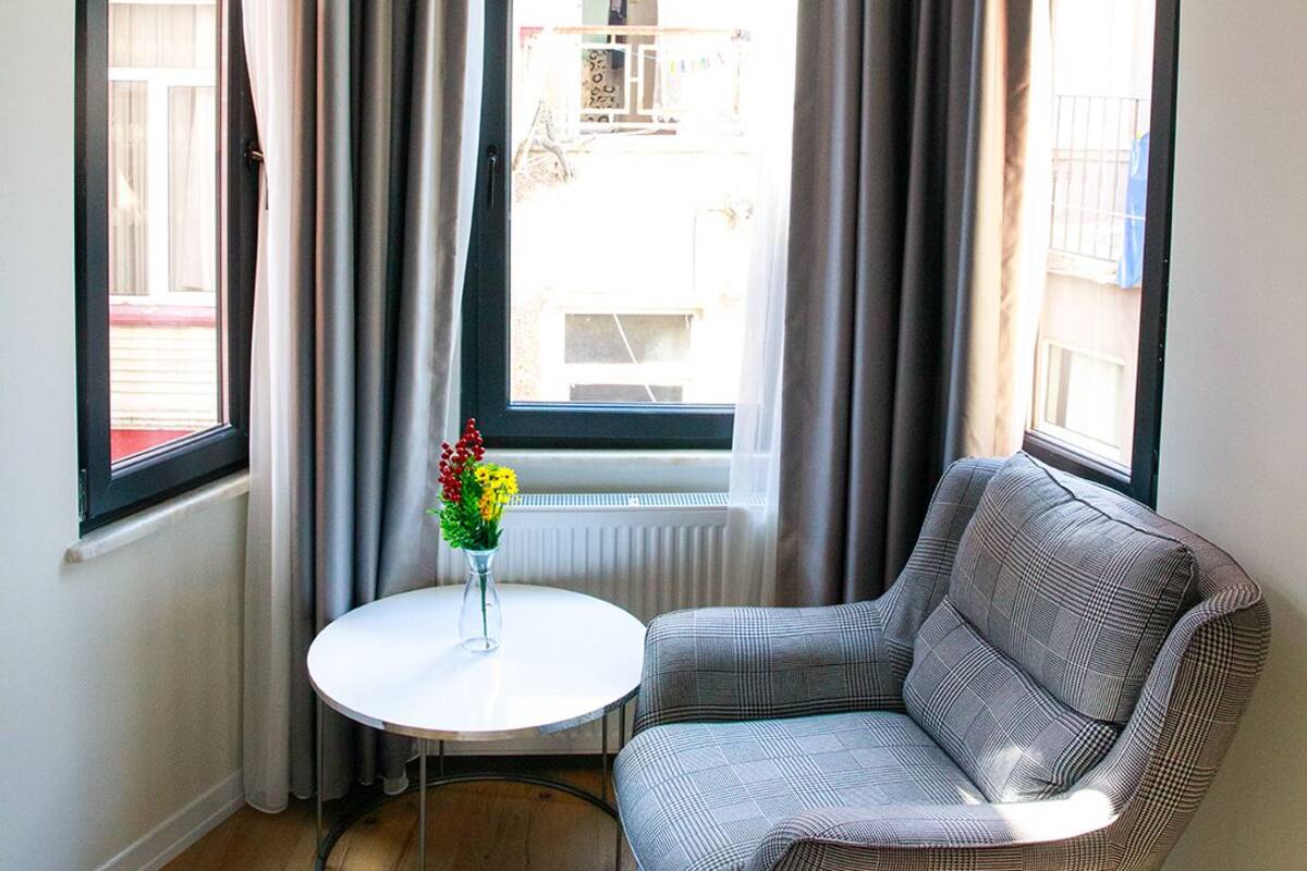 A corner room with a grey armchair, a small round table with a vase of flowers, and a window with curtains.