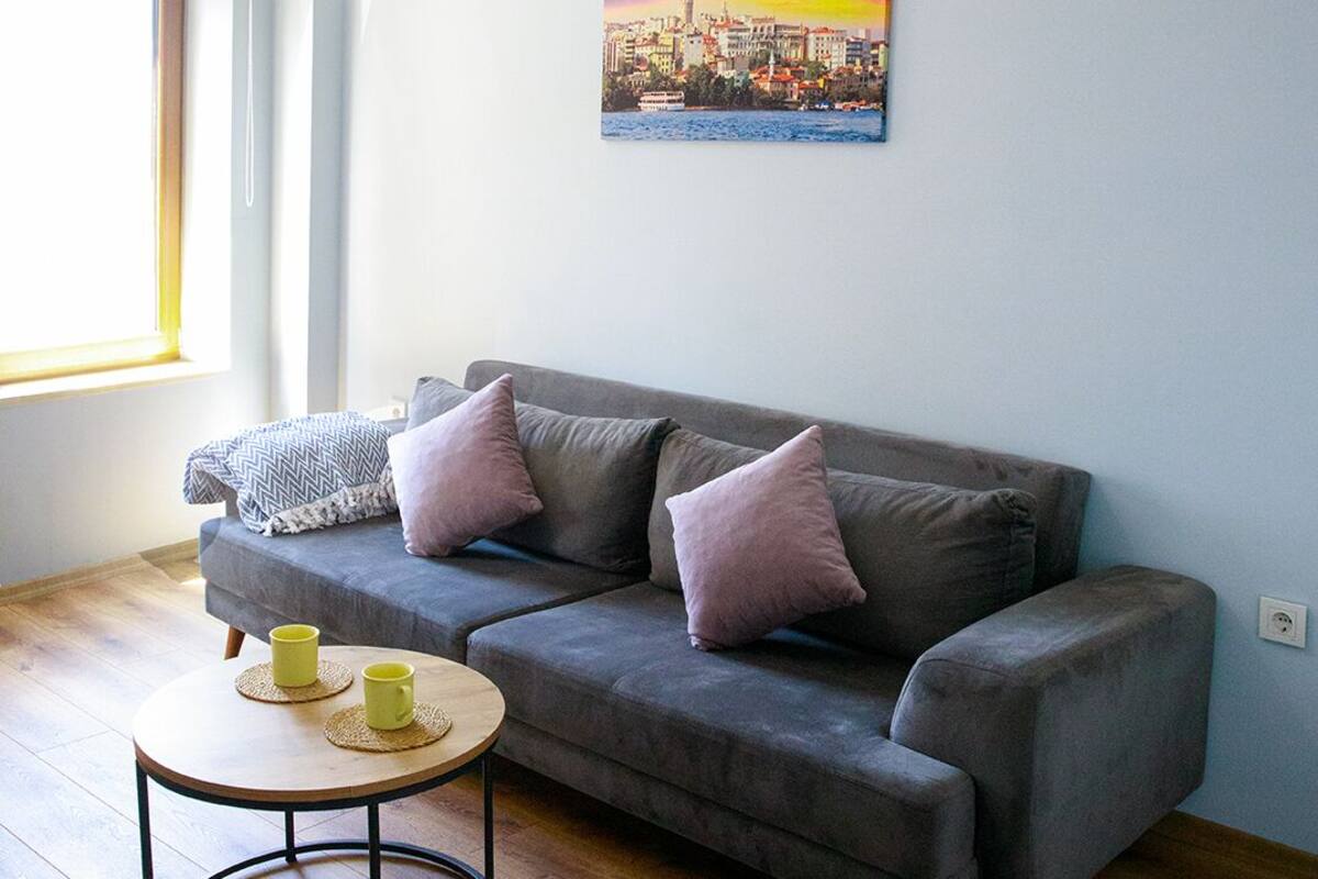 A living room with a grey sofa, two cushions, a round coffee table with two yellow mugs, and a cityscape painting on the wall.