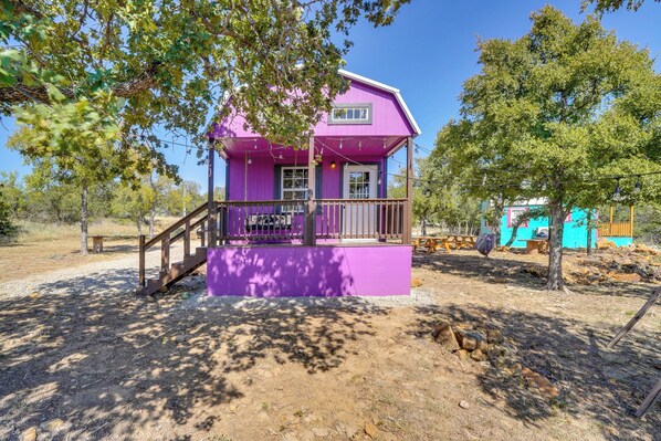 A purple house with a porch and a small deck, surrounded by trees and a dirt ground.