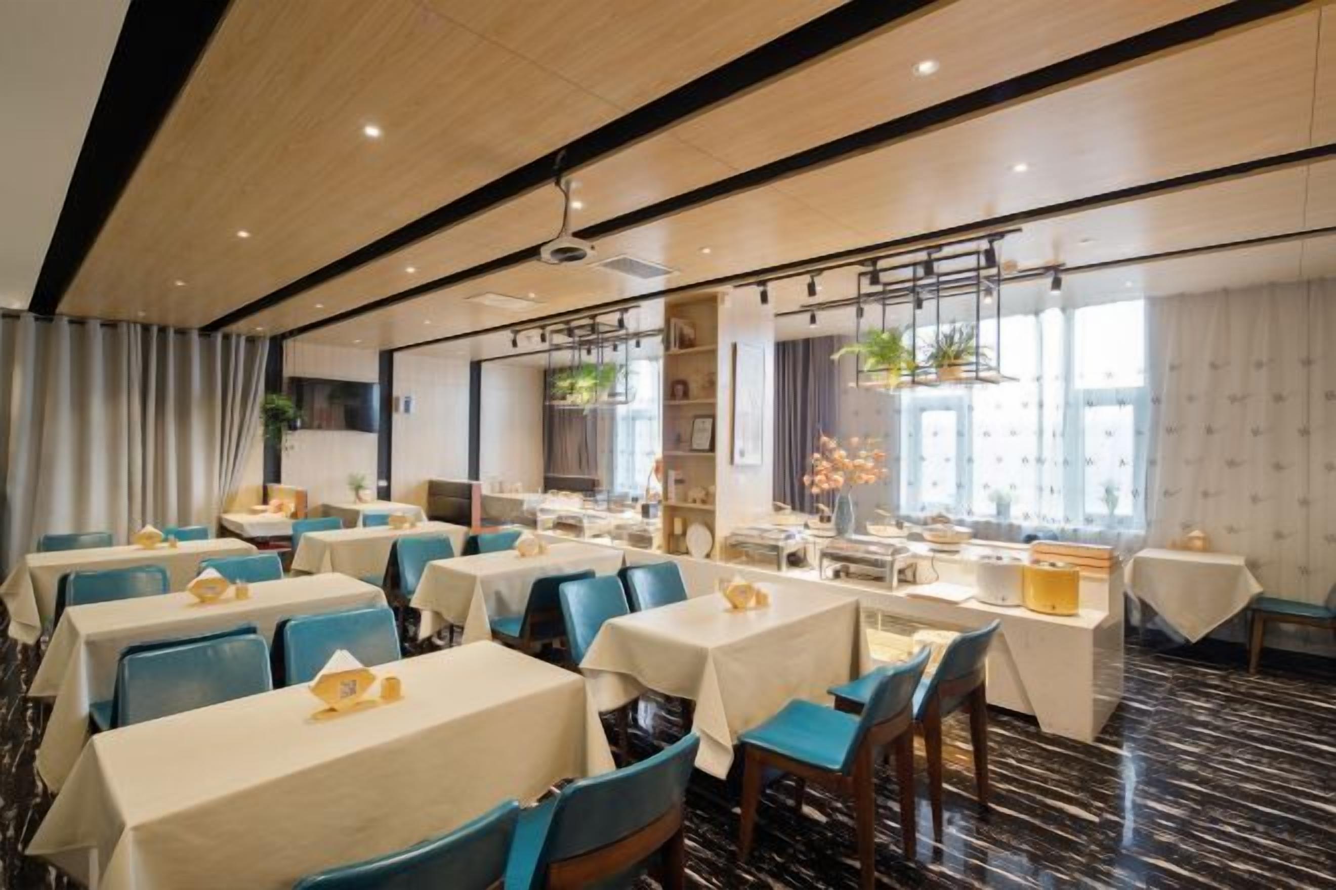 Uma sala de jantar de hotel com várias mesas e cadeiras, um ventilador de teto e janelas amplas.