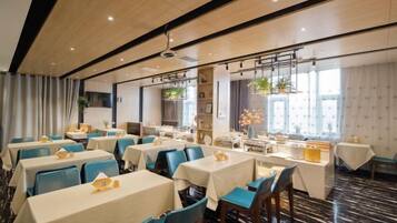 Uma sala de jantar de hotel com várias mesas e cadeiras, um ventilador de teto e janelas amplas.