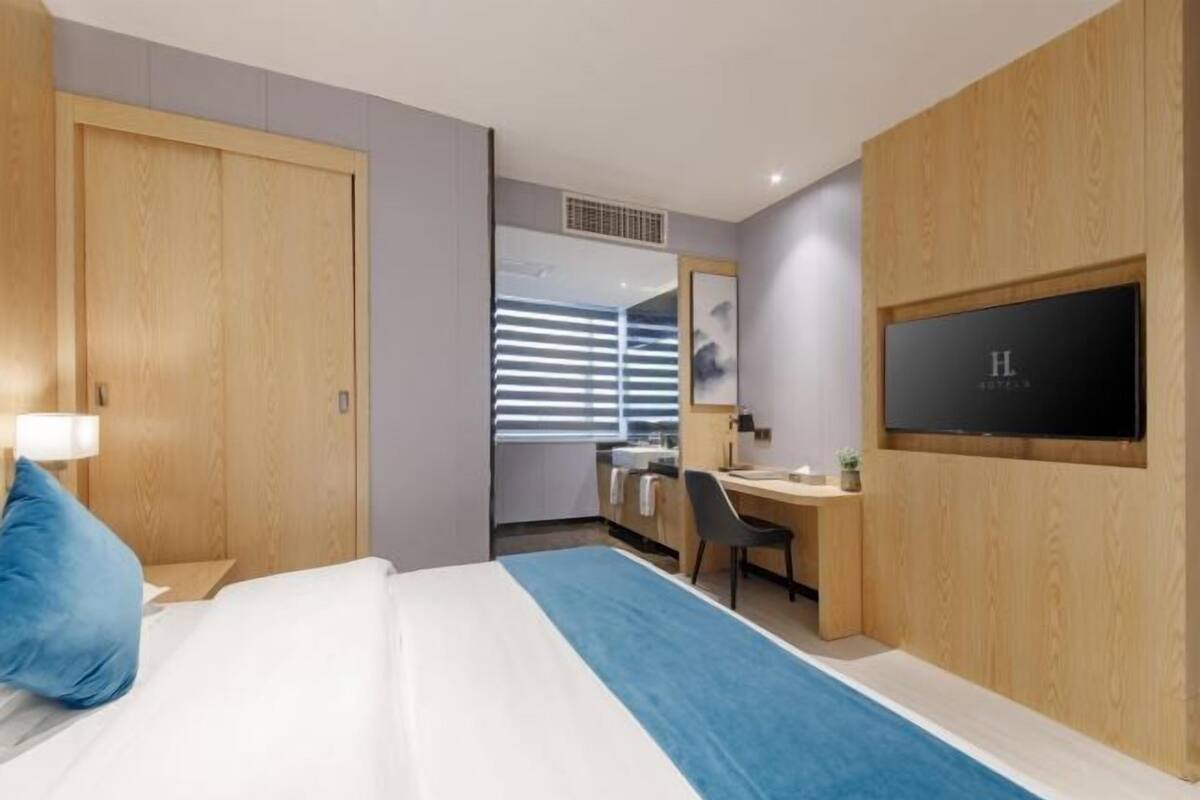 A hotel room with a bed, a desk, a chair, a TV, and a window with blinds.
