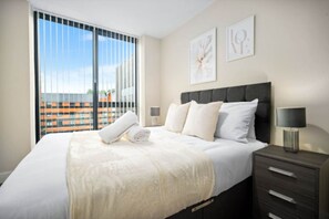 A modern hotel room with a large bed, a nightstand, a lamp, and a view of a cityscape through the window. - Two Bed Apartment - Smart TV - WIFI - Jewellery Quarter (Birmingham)
