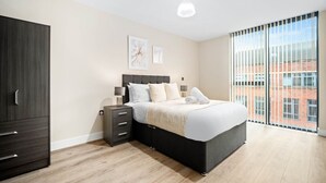 A modern bedroom with a large bed, a dark wardrobe, a bedside table with lamps, and a large window with a city view.