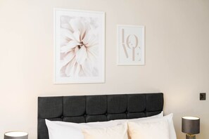 Un dormitorio con un cabecero tapizado en gris oscuro, una obra de arte floral enmarcada y una pieza de arte con palabras en la pared.