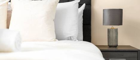 A neatly made bed with white bedding and pillows, a dark headboard, and a bedside table with a lamp.