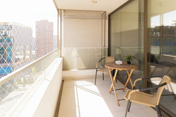 Een balkon met een glazen deur, twee rieten stoelen, een klein rond tafeltje en een bloempot.