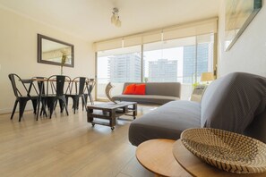 Een moderne woonkamer met een grijze bank, een eettafel met stoelen en een groot raam met uitzicht op de stad.