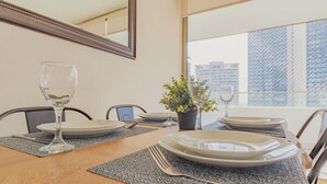 A dining table set with plates, glasses, and cutlery, a potted plant, and a view of buildings through a window. - Charming 2-bedroom apartment with AC, WiFi in Parque Arauco (Vitacura)