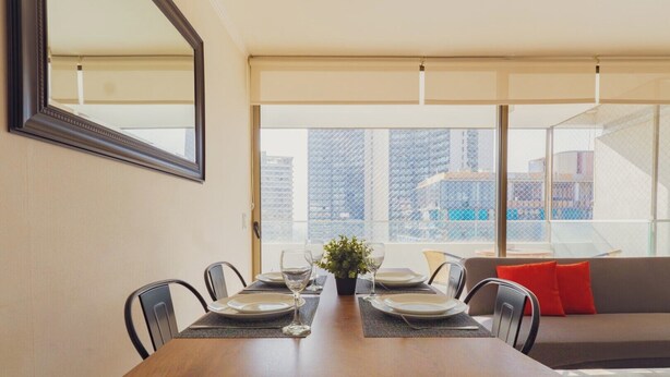 A dining area with a table set for four, a large window with a view of buildings, and a sofa with red cushions. - Charming 2-bedroom apartment with AC, WiFi in Parque Arauco (Vitacura)