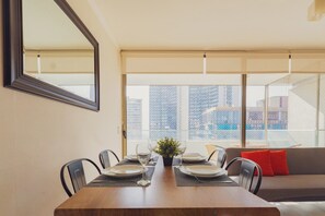 Un comedor con una mesa dispuesta para cuatro, un ventanal con vistas a edificios, y un sofá con cojines rojos.