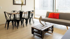 A modern living room with a dining table, chairs, a sofa with orange cushions, and a large window with a city view. - Charming 2-bedroom apartment with AC, WiFi in Parque Arauco (Vitacura)