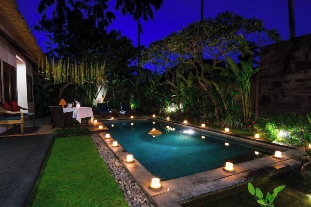 A nighttime swimming pool with candles along the edge, surrounded by lush greenery and a thatched-roof building.