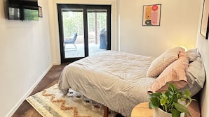 Un dormitorio con cama, una mesita con una planta, un televisor montado en la pared y una puerta corrediza de vidrio que da a un espacio exterior.