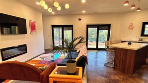Una sala de estar moderna con un sofá, una mesa de centro y un televisor montado en la pared. Se ven grandes ventanales y una zona de comedor al fondo.
