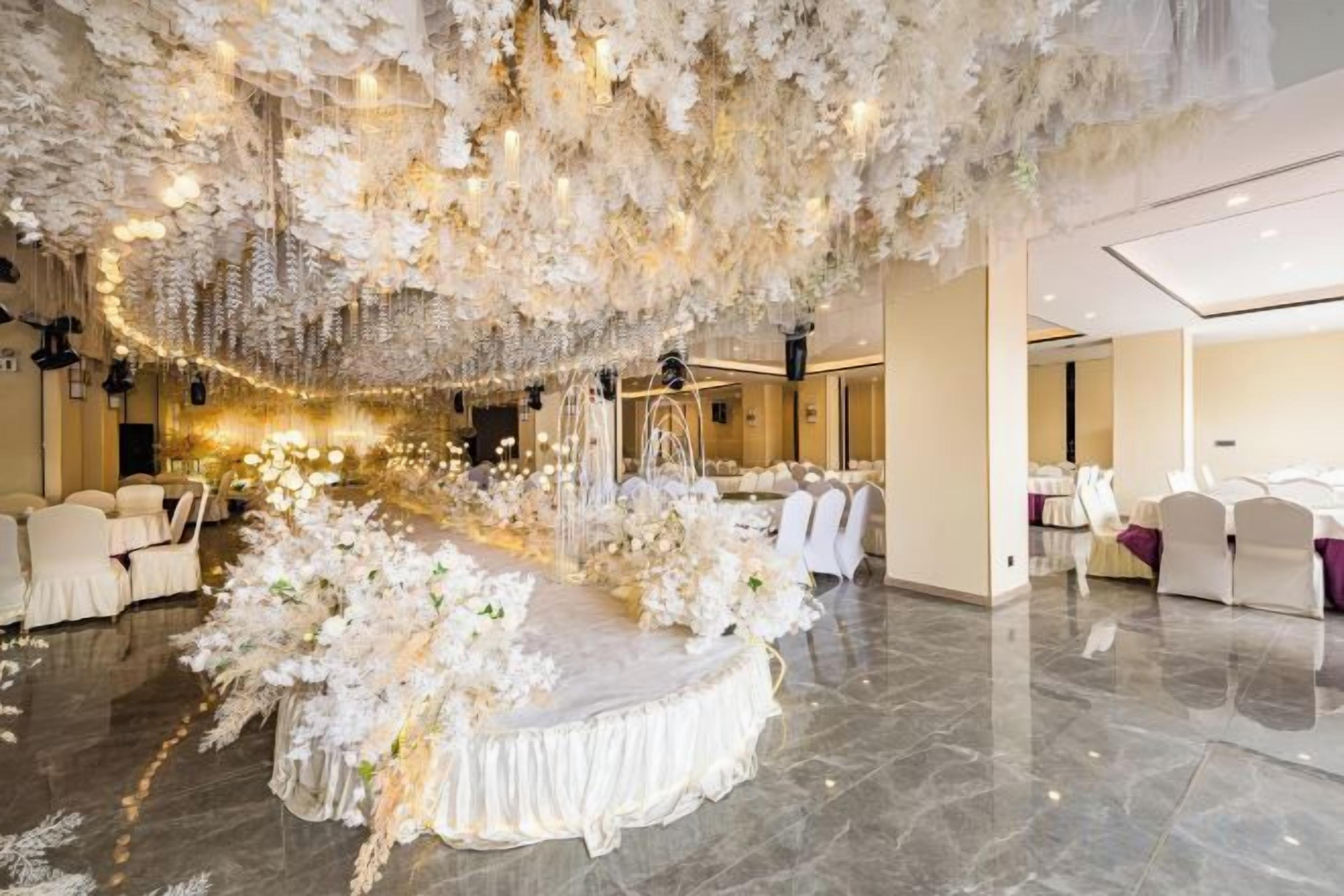 A banquet hall with a round table adorned with white flowers and a tiered floral arrangement.