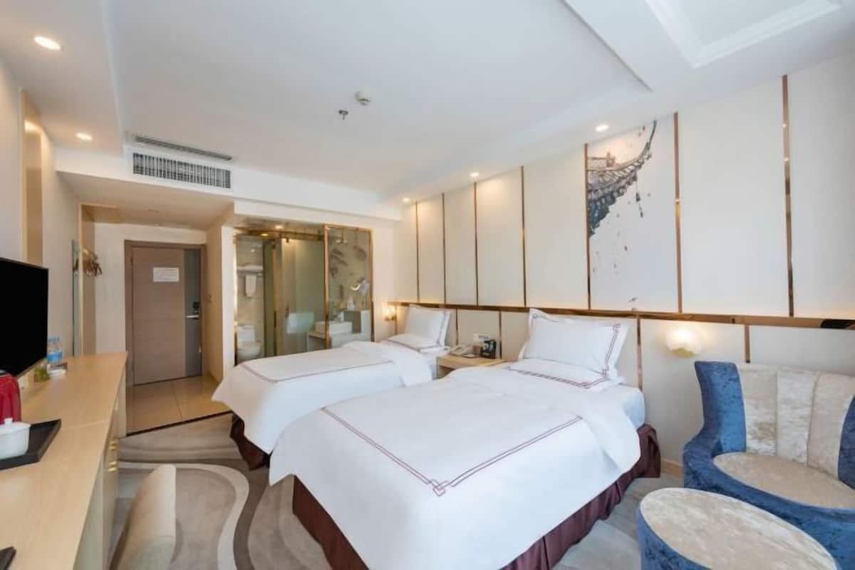 Habitación de hotel con dos camas, un escritorio, un televisor y baño visible a través de una puerta de cristal.