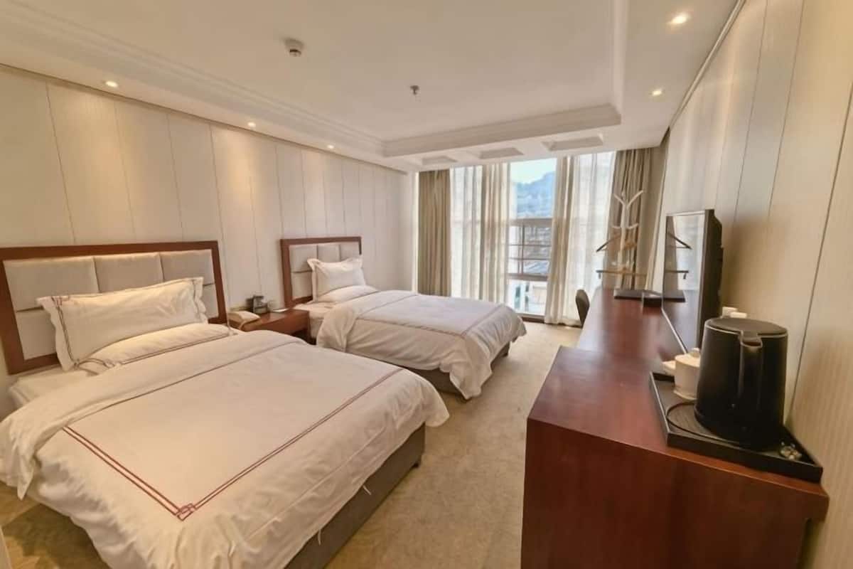 Habitación de hotel con dos camas, un escritorio con cafetera y vistas a la ciudad a través de amplias ventanas.