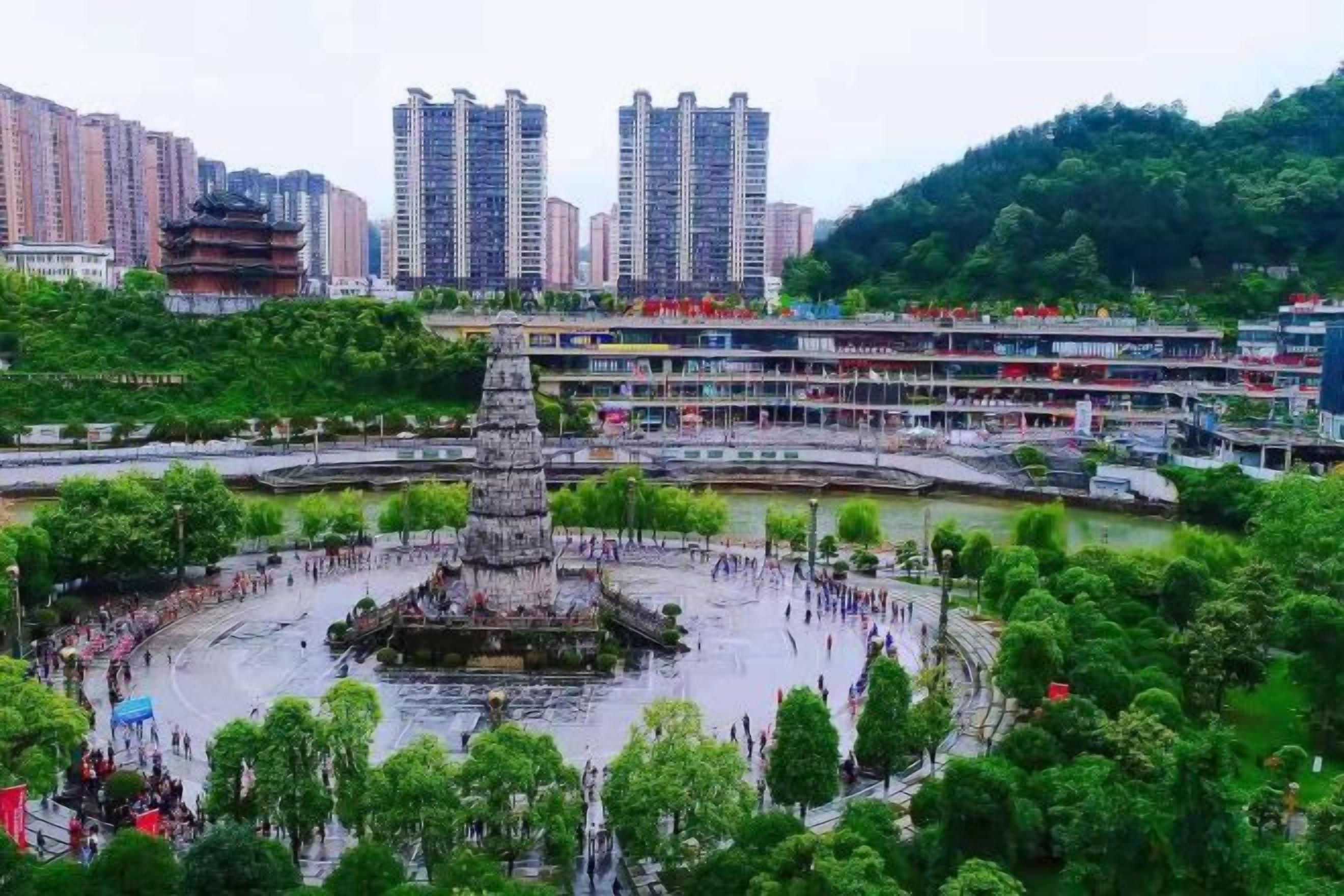 A public square with a central monument, surrounded by greenery and buildings.