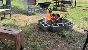 A backyard with a fire pit surrounded by concrete blocks, a wheelbarrow, a grill, and a chair.