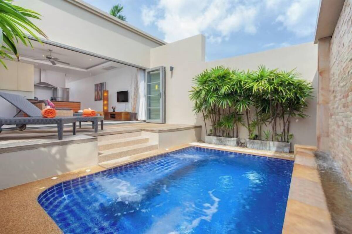 Un área de piscina moderna al aire libre con un jacuzzi, sillones de descanso y un jardín bien cuidado con plantas altas.