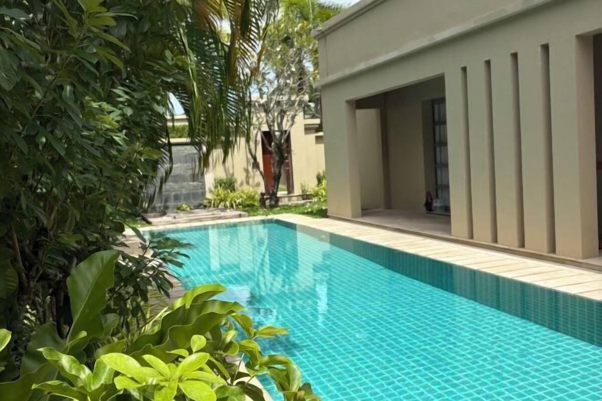 Una piscina con bordes de azulejos, rodeada de exuberante vegetación y un edificio moderno con un amplio ventanal.