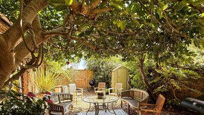 Een terras met een ronde glazen tafel, omringd door houten stoelen, een kleine schuur en een barbecue.