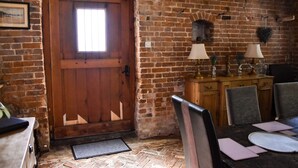 Een rustieke kamer met een houten deur, bakstenen muren, een eettafel met stoelen en een klein kastje met lampen.