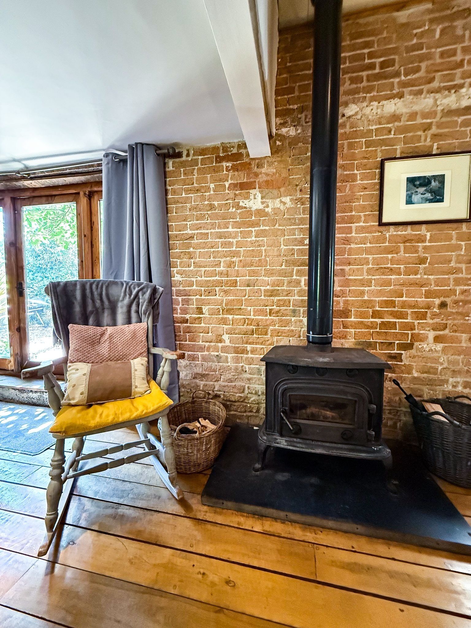 Une pièce rustique avec un mur en briques, une chaise en bois avec des coussins, un poêle à bois noir et un panier en osier posé sur le sol.