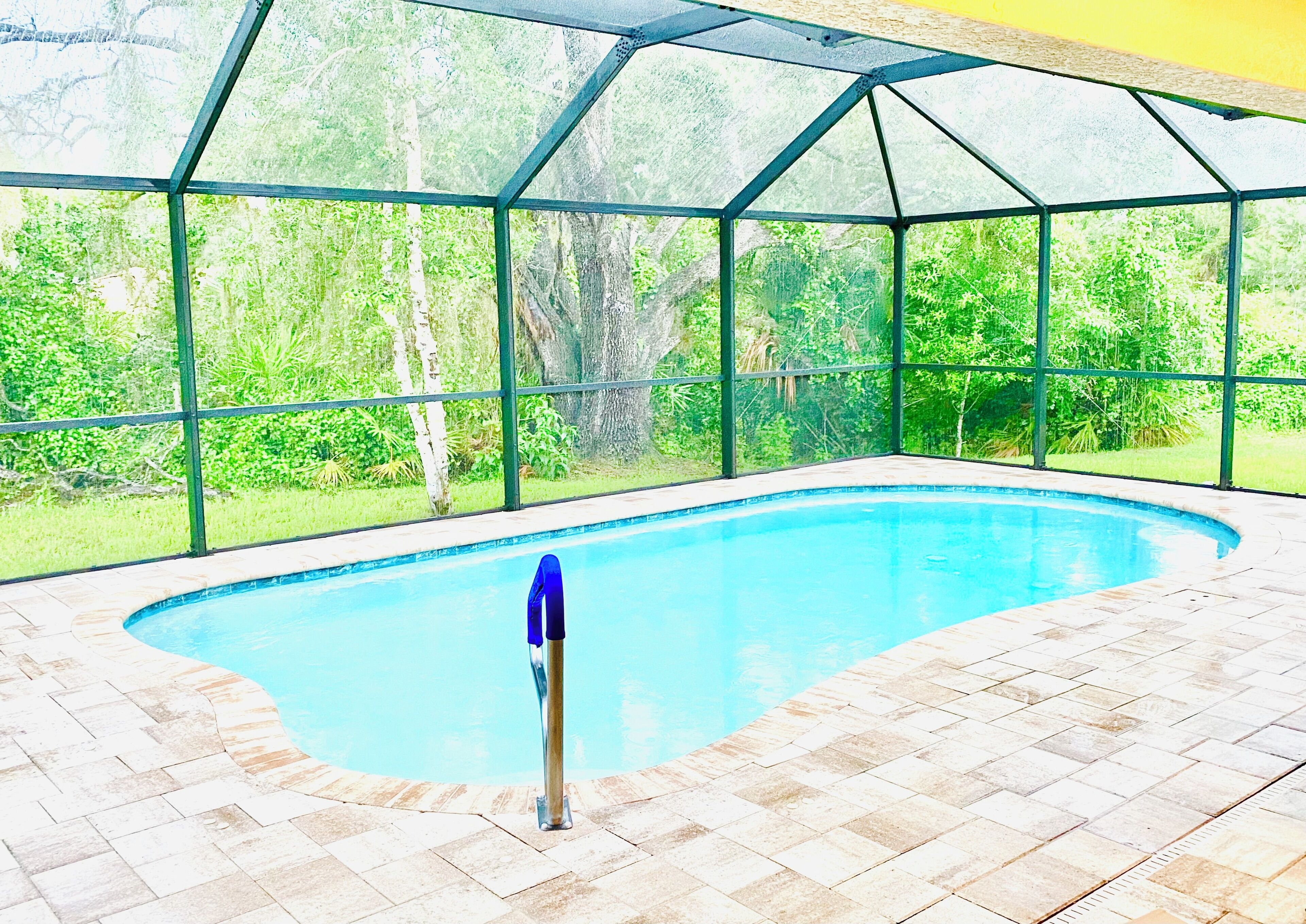 An indoor swimming pool with a glass enclosure, surrounded by a tiled floor and a view of greenery outside.
