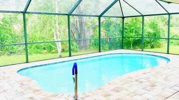An indoor swimming pool with a glass enclosure, surrounded by a tiled floor and a view of greenery outside.