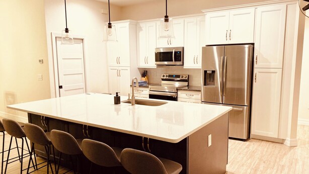 A modern kitchen with white cabinets, stainless steel appliances, a large island with bar stools, pendant lighting, and a wooden floor.
