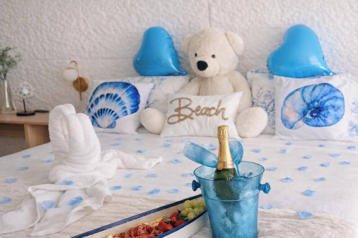 Una cama con una colcha blanca y azul estampada con flores, un peluche de oso y almohadas azules con forma de corazón.