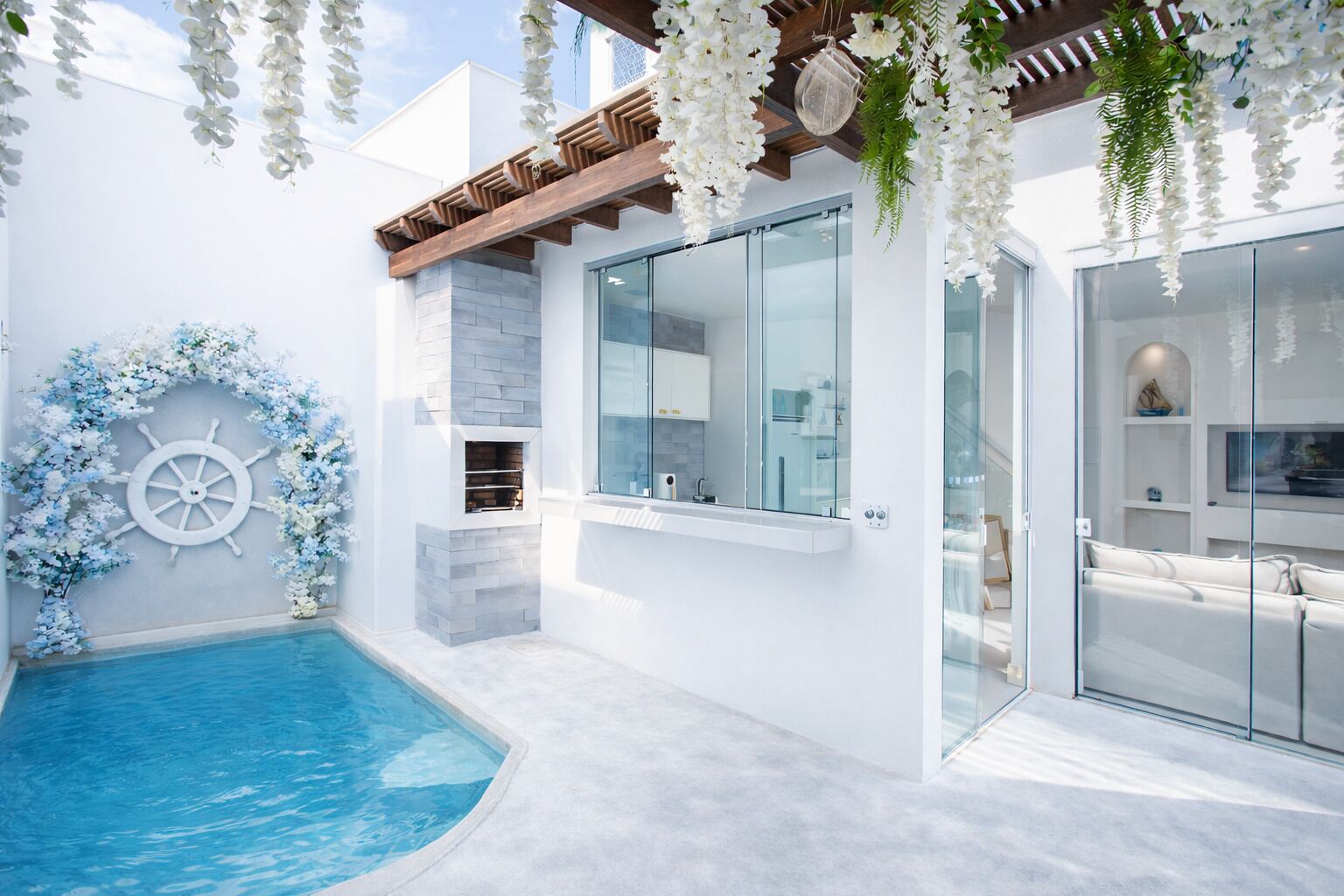 A modern indoor pool area with a stone fireplace, glass shower enclosure, and a nautical-themed decor with a ship's wheel and white flowers.