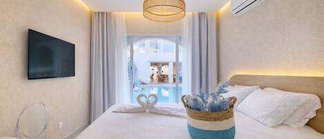 Quarto de hotel moderno com uma cama grande, uma TV fixada na parede, uma luminária no teto e vista para a área da piscina.