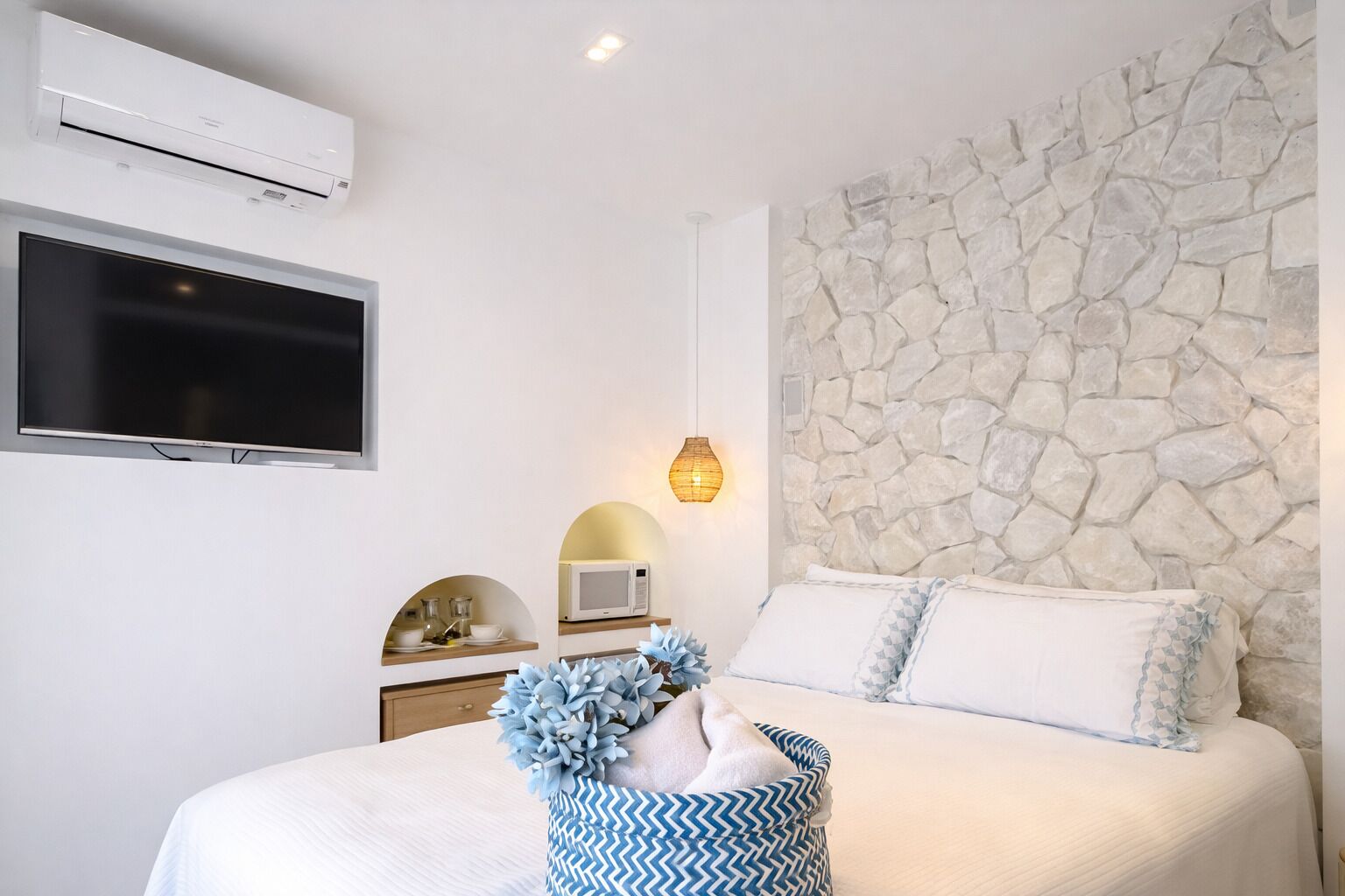 A bedroom with a stone wall, a flat-screen TV, a microwave, and a bedside table with a vase of flowers.