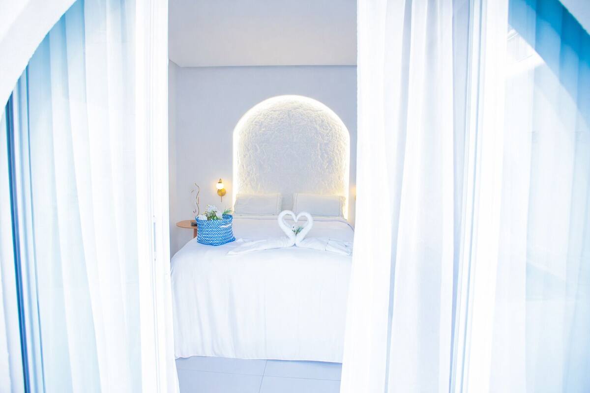 Un dormitorio con una cama grande, una decoración con forma de corazón sobre la cama y una cesta blanca y azul estampada en la mesita de noche.