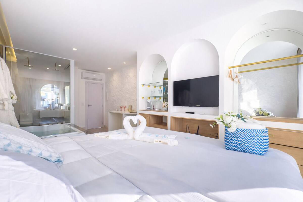 Una habitación de hotel moderna con una cama grande, televisión, tocador con espejo y baño visible a través de una puerta de vidrio.