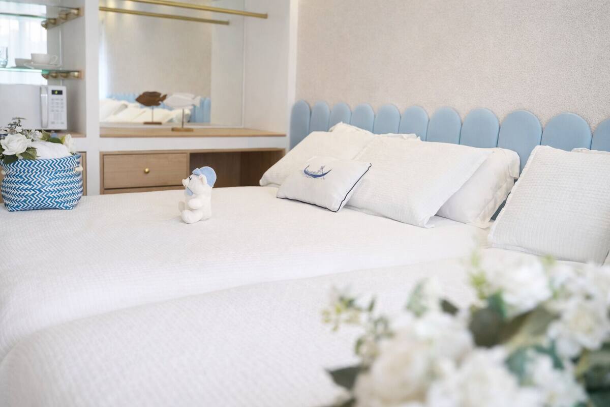 Una cama bien hecha con ropa blanca y almohadas, una mesita de noche con una cesta azul y blanca estampada, y un teléfono de pared.