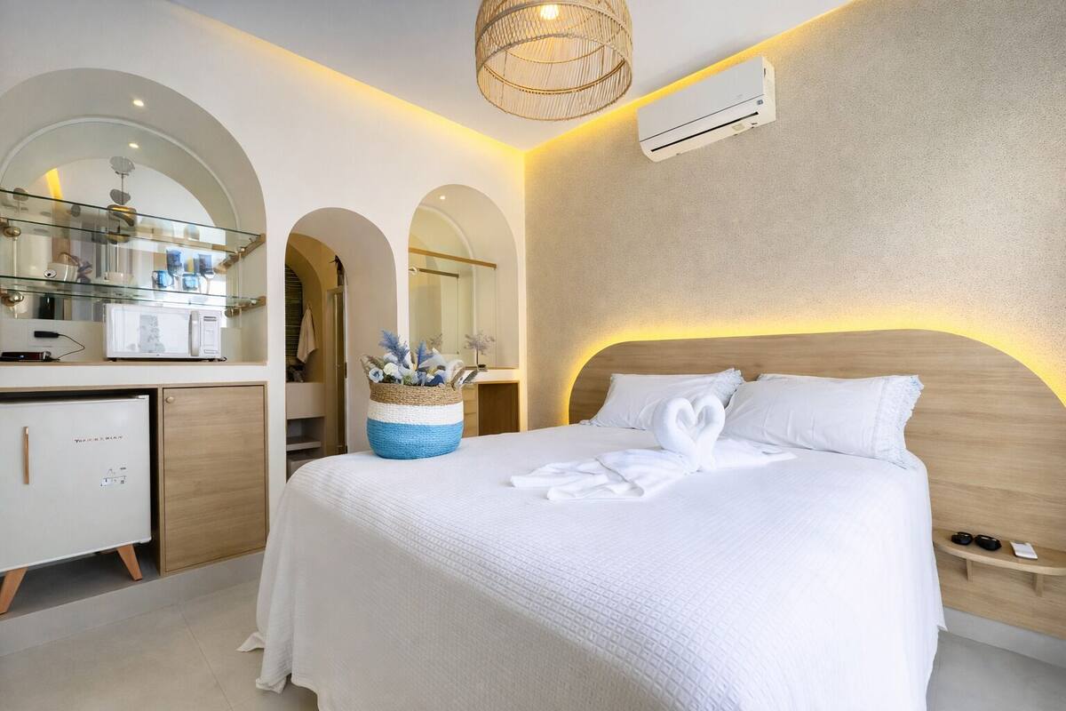 Una habitación de hotel moderna con una cama grande, cabecera de madera, aire acondicionado integrado y un baño con mampara de vidrio, tocador y espejo.